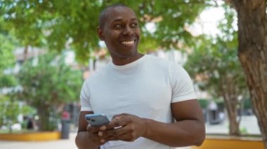 Çevresi yemyeşil ve şehir manzarasıyla çevrili, şehir parkında akıllı telefon kullanan gülümseyen adam, açık havanın tadını çıkarıyor..