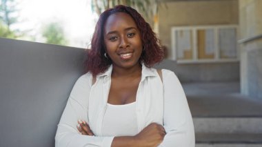 Genç Afrikalı Amerikalı bir kadın şehir sokaklarında dikiliyor, kameraya güvenle gülümsüyor, kollarını kavuşturmuş..