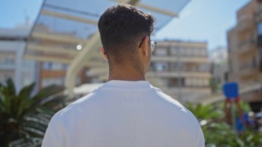 Yakışıklı, genç, İspanyol bir adam açık havada, şehir parkında, aydınlık bir günde etrafı binalarla çevrili duruyor..