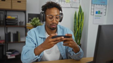 Sakallı, genç bir Afrikalı Amerikalı telefonuyla video oyunu oynuyor. Ofis ortamında kulaklık takıyor..