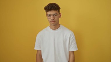 Yakışıklı, gözlüklü ve beyaz tişörtlü genç bir adam izole edilmiş sarı bir arka plana karşı duruyor. Basit ama büyüleyici bir portrede kendine güven ve tarz yayıyor..
