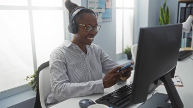 Genç, Afrikalı, Amerikalı, ofiste gülümseyen, kulaklık takan ve elinde cep telefonuyla bilgisayar başında çalışan bir kadın.