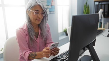 İş yerinde arka planda bilgisayarla cep telefonu kullanan bir kadın, ofis ortamında modern teknoloji kullanımını gösteriyor