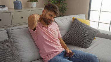 Oturma odasındaki koltukta oturan sakallı genç bir adam modern bir dairede boynunu tutarken telefonunu tutuyor.