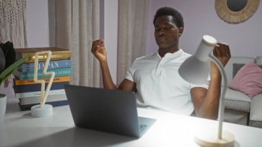 Genç bir adam evinde, oturma odasında dizüstü bilgisayarla oturmuş meditasyon yapıyor.