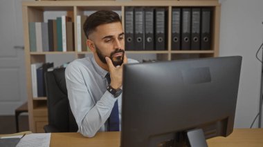 Ofiste bilgisayar başında çalışan sakallı genç adam, odaklanmış ve düşünceli, raflar ve belgelerle sıradan çalışma ortamı, profesyonel atmosferi vurgulayan kapalı bir portre..