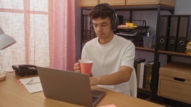Kulaklık takan, elinde kupa tutan, rafları ve arka planda çeşitli belgeleri olan bir ofiste dizüstü bilgisayarla çalışan İspanyol bir genç..