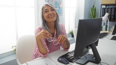 Parlak ofis çalışma odasında elinde bilgisayar ve gözlüklerle masasına gülümseyen yaşlı gri saçlı kadın.