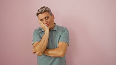 Kollarını kavuşturmuş yakışıklı genç bir adam ve soyutlanmış pembe bir arka plana karşı düşünceli bir ifade.