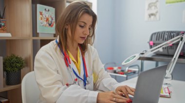 Genç ve çekici sarışın bir kadın veteriner kliniğinde dizüstü bilgisayarla çalışırken steteskopla laboratuvar önlüğü giyiyor..