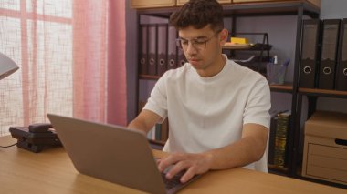 İyi organize edilmiş bir ofis ortamında dizüstü bilgisayar üzerinde çalışan genç bir İspanyol adam, konsantre olarak görevine odaklanıyor..