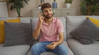 Genç İspanyol bir adam kulaklık takıyor ve elinde akıllı bir telefon, gri bir kanepede oturuyor. Oturma odasında, renkli yastıklar ve bitkilerle..