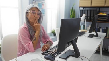 Gri saçlı yaşlı bir kadın ofis ortamında bilgisayar başında çalışırken sessizlik jesti yapıyor.