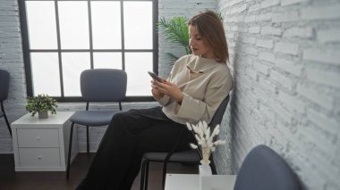 Çekici, genç, sarışın kadın modern bir bekleme odasında oturuyor. Telefonunu bitkilerle ve basit mobilyalarla kullanıyor..