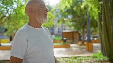 Olgun bir adam güneşli bir günde arka planda yeşillikle şehir parkında duruyor, beyaz gömlek giyiyor ve gri sakallı..