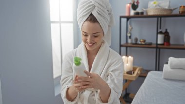 Kaplıcaya gülümseyen bir kadın kafasında havlu, arka planında mumlar ve raflarla sağlık merkezinde bornoz takıyor.