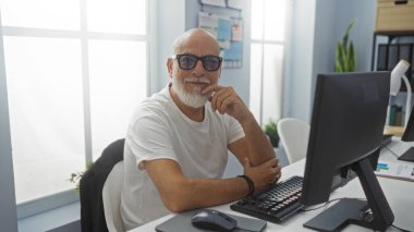 Gri saçlı ve sakallı yakışıklı bir adam ofiste oturmuş kameraya bakıyor.