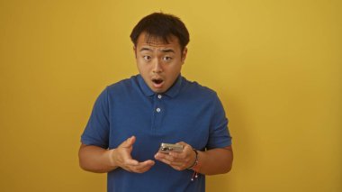 Mavi gömlekli çekici Çinli bir adam, izole edilmiş sarı arka plan duvarına akıllı telefon tutarken şaşırmış görünüyor..