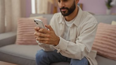 Sakallı genç İspanyol bir adam pembe yastıklı ve bej perdeli rahat bir oturma odasında otururken akıllı telefon kullanıyor.