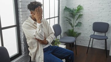 Kapalı bir bekleme odasında sakallı, başı dertteyken sandalyede oturan Afrikalı Amerikalı genç bir adam.