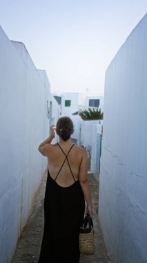 Aile, Lanzarote 'de dar, beyaz yıkanmış bir sokakta yürüyen siyah elbiseli ve elinde sepeti olan bir kadın açık havada canlı İspanyol deniz kenarı manzarası sergiliyor.