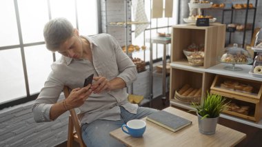 Genç adam, fırında cep telefonuyla kahve fincanının fotoğrafını çekiyor. Etrafı çeşitli fırınlarla çevrili. Geniş pencerelerden de doğal ışık.