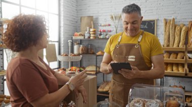 Kadın ve erkek, pastanede hamur işleri ile etkileşime giriyor ve adam dijital tablete danışırken önlük takıyor.