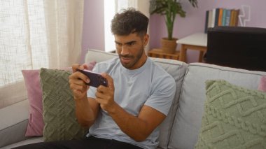 Genç İspanyol adam rahat oturma odasında pastel dekorlu ve pelüş minderli akıllı telefonla oynuyor.