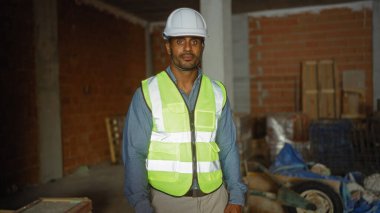 Başlık ve güvenlik yeleği giyen yakışıklı Afro-Amerikan erkeği inşaat malzemelerinin geçmişine sahip aktif bir şantiyede güvenle duruyor.