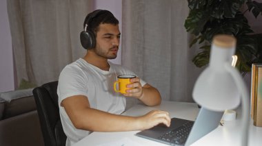 Evde dizüstü bilgisayar kullanan, elinde sarı kupa olan ve oturma odasında kulaklık takan genç bir İspanyol.