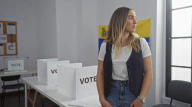 Venezuela 'da bir seçim koleji odasında oy verme kabinleri ve arka planında Venezuela bayrağı olan güzel sarışın bir kadın..