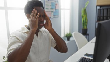 Genç, Afro-Afrikalı, ofisteki Amerikalı bir adam başını tutuyor, modern bir iç mekan geçmişi olan bir bilgisayarın yanında stresli görünüyor..