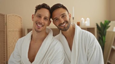 Sıcak bir sağlık merkezinde birlikte gülümseyen bornozlu iki adam, samimi bir spa ortamında sevgi ve rahatlığın özünü yakalıyorlar.