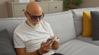 Gri sakallı bir adam, oturma odasında gri bir koltukta oturuyor, ilaç şişesini ve akıllı telefonunu kontrol ediyor, modern dekorla kapalı bir yerde, gözlük takıyor ve günlük beyaz tişört giyiyor.