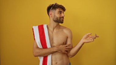 Genç adam, omzunda havluyla sarı bir arka plana karşı kendine güvenen bir gülümseme ile yan tarafı işaret ediyor.