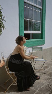 İspanyol kadın dışarıda bir otel restoranının terasında oturuyor, güneşli havanın keyfini çıkarırken yeşil pencere çerçevesinin yanında siyah bir elbise giyiyor..