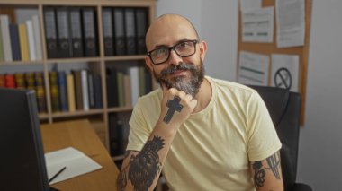 Ofis odasında sakallı, kolunda ve elinin altında kitaplık ve dövmelerle oturan İspanyol bir adam.