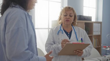 Orta yaşlı kadın doktor bir hastayla konuşurken klinikte notlar alıyor..