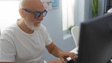 Gri saçlı ve sakallı kıdemli bir adam ofis ortamında bilgisayarın başında çalışıyor. İçerideki işine odaklanmış.