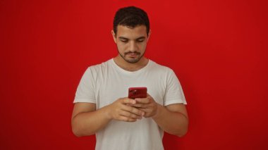 Yakışıklı İspanyol genç adam izole edilmiş kırmızı arka plan üzerinde akıllı telefon kullanıyor.