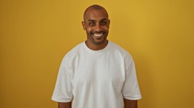 Genç Afro-Amerikalı adam, izole edilmiş beyaz tişörtlü sarı duvarın üzerinden güvenle gülümsüyor.
