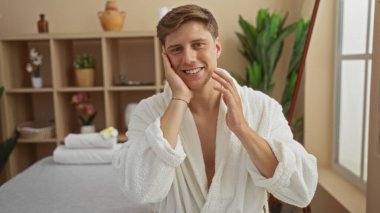 Beyaz bir bornoz giyen, gülümseyen ve yüzüne dokunan genç bir adam. Etrafı bitkiler ve zenginlik dekoru ile çevrili..