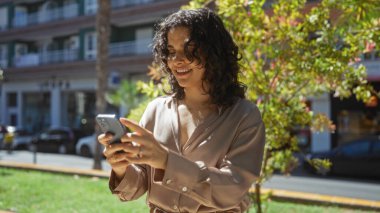 Şehir parkında akıllı telefon kullanan güzel genç bir kadın mutluluğunu gösteriyor ve şehir yaşamını kucaklıyor..