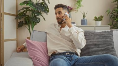 Genç, yakışıklı, İspanyol bir adam rahat, şık bir oturma odasında bir koltukta oturuyor, cep telefonuyla konuşuyor, iç mekan bitkileri, yastıklar ve modern dekorlarla çevrili..