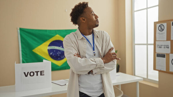 Красивый молодой африканский американец с бородой стоит со скрещенными руками в помещении бразильского избирательного колледжа, с флагом, кабинкой для голосования и информационным баннером.