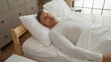 Orta yaşlı bir adam, beyaz bir yatakta, rahat bir yatak odasında uyur. Pencereden süzülen yumuşak doğal ışık.