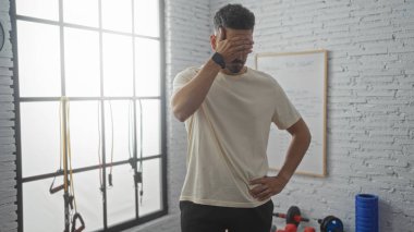 Egzersiz bantları ve ağırlıklarıyla spor salonunda duran yakışıklı genç adam, düşünceli ya da yorgun görünüyor..