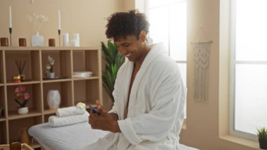 Huzurlu spa odasında telefona gülümseyen bornozlu genç adam. Sıcak aydınlatma ve modern dekorla.