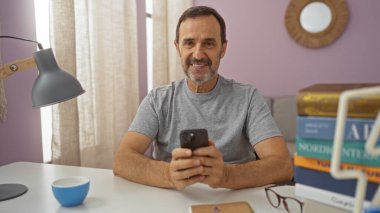 Yakışıklı, İspanyol asıllı bir adam evde oturma odasında oturuyor, elinde gülümsemeli bir akıllı telefon, etrafı kitaplarla çevrili ve beyaz bir masada bir lamba tutuyor..