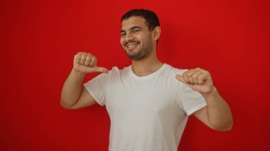 Beyaz gömlekli, izole edilmiş kırmızı arka planda güvenle gülümseyen İspanyol genç adam iki başparmağıyla kendini işaret ediyor.